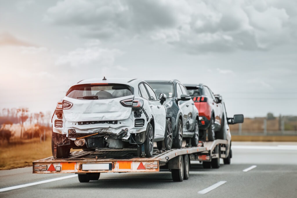 White SUV with rear-end damage on a tow trailer, followed by other damaged cars on a highway transport truck under a cloudy sky, road ahead.