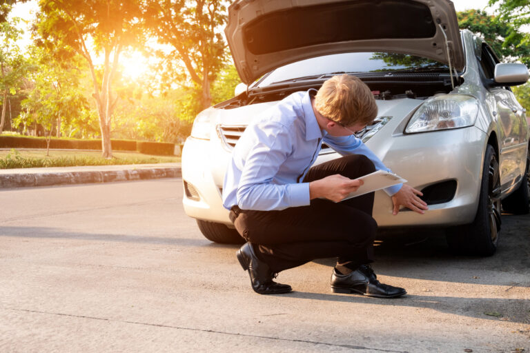 Man kneeling by a silver car with its hood open, examining a clipboard on a sunny roadside repair. Used as a roadside assistance scene.