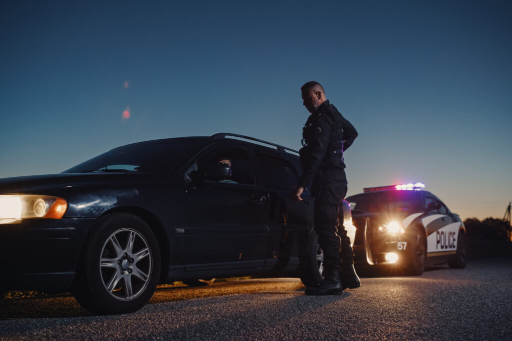 Officer in tactical gear beside a dark sedan during dusk with flashing police lights in the background, outside a traffic-stop scene.