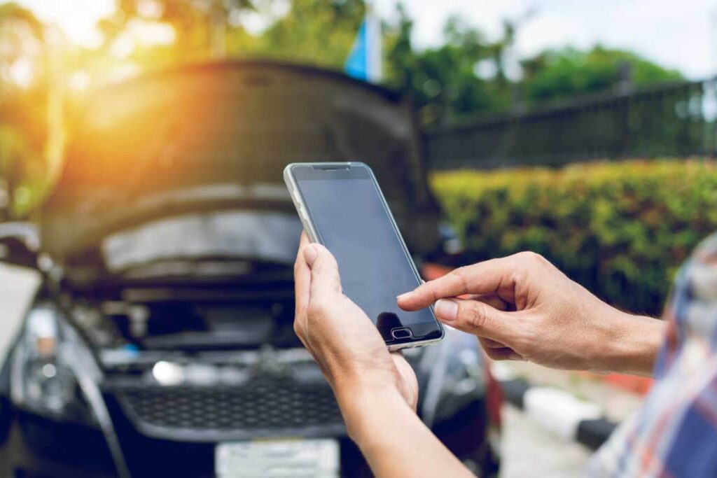 Person holding a smartphone beside a parked car outdoors, sunlight on the left.
